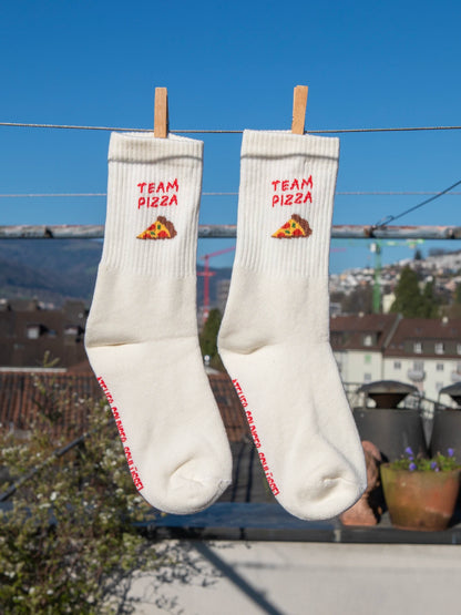 Zwei ecru farbenen Sportsocken hängen an einer Wäscheleine. Im Hintergrund sieht man unscharf Häuser, Hügel und einen blauen Himmel. Im Bündchen der beiden Socken ist der Schriftzug Team Pizza sowie ein Stück Pizza gestrickt.