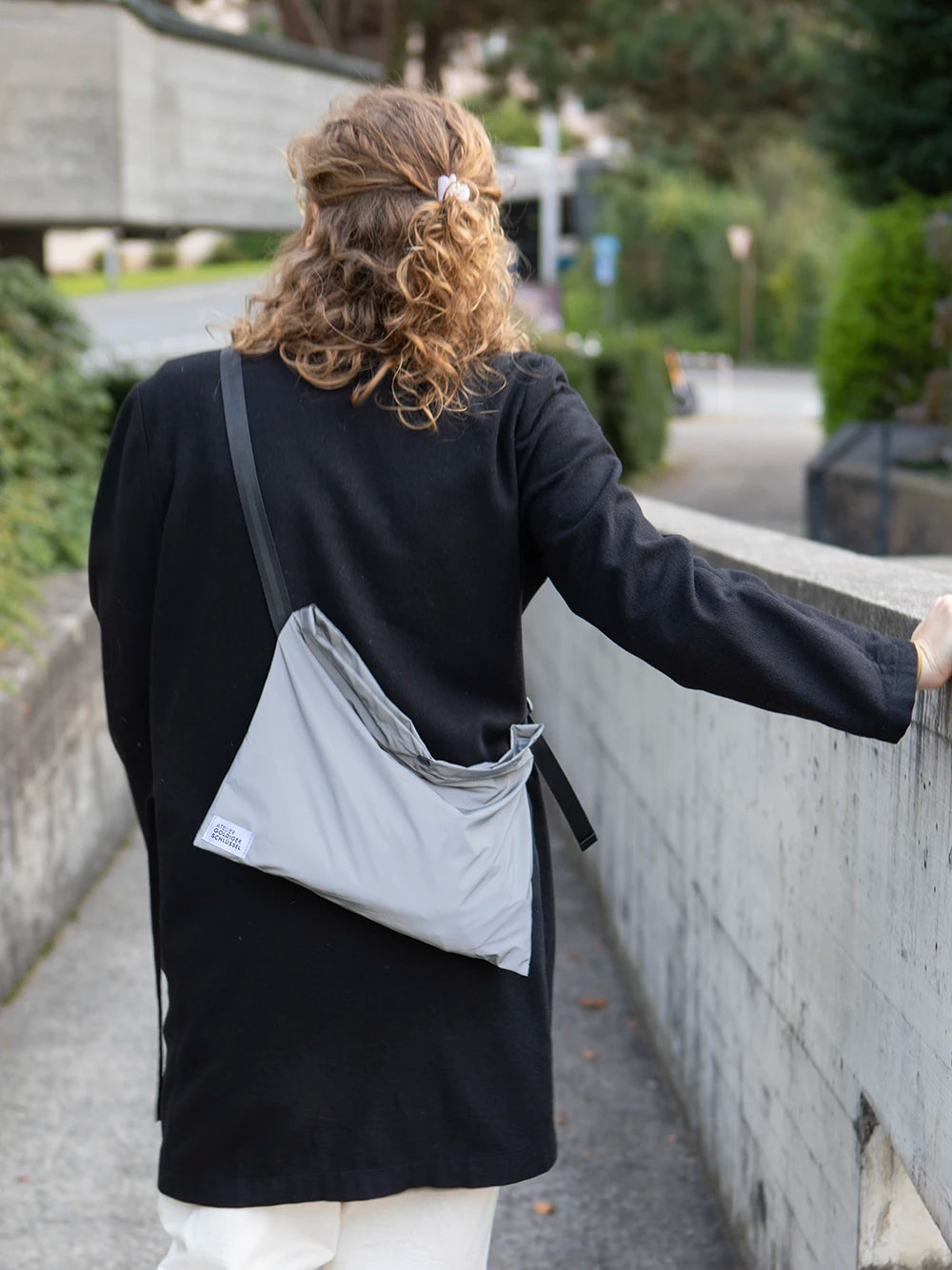 Rückseite des Oberkörpers einer Person mit blonden Locken und einer schwarzen Jacke welche eine reflektierende Umhängetasche mit dem Label «Atelier Goldiger Schlüssel» trägt.