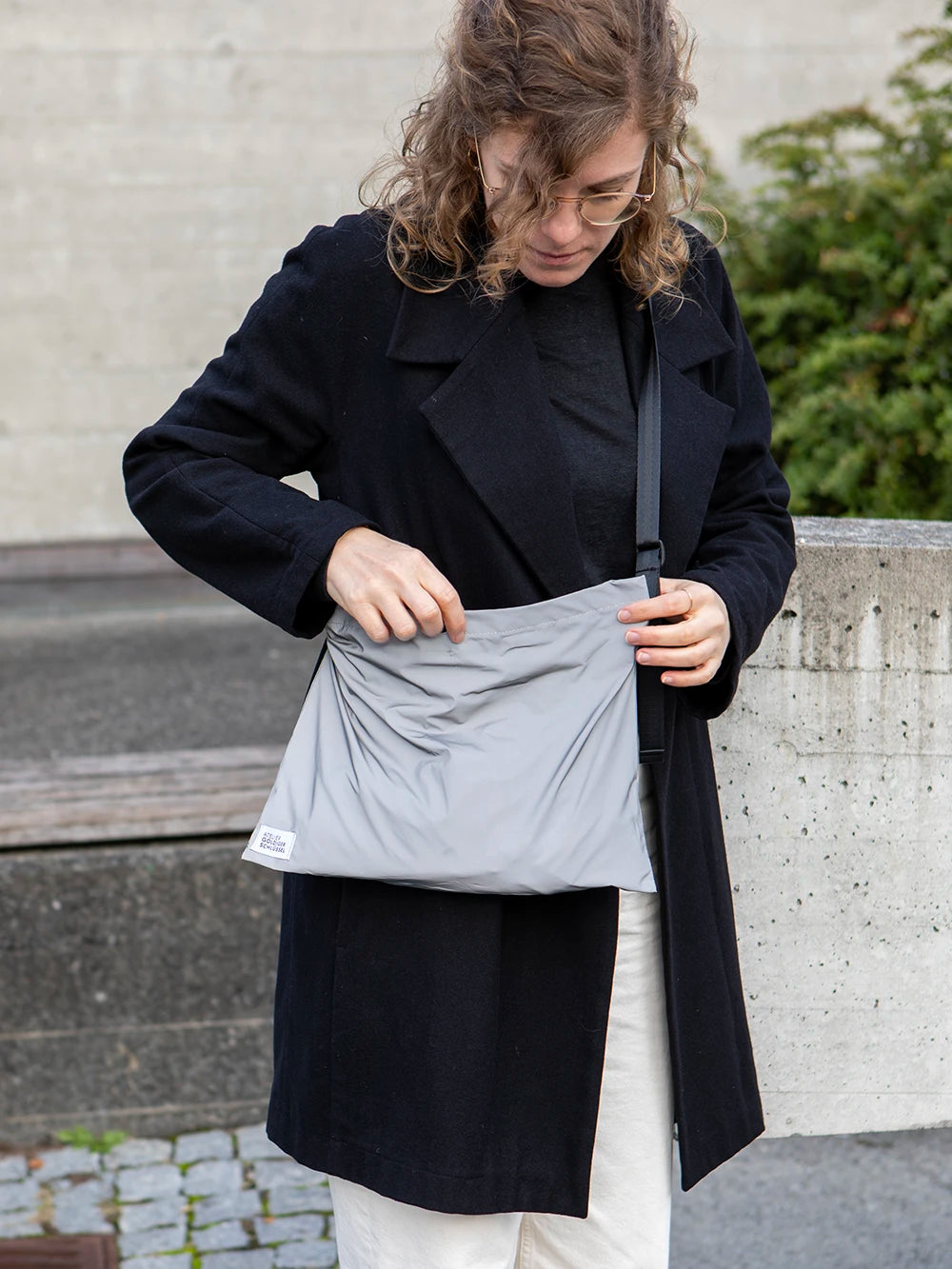 Frau mit blonden Locken und einer schwarzen Jacke trägt eine reflektierende Umhängetasche mit dem Label «Atelier Goldiger Schlüssel».