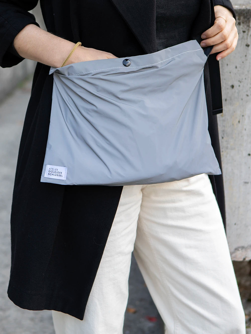 Person mit schwarzer Jacke und nature farbenen Hosen trägt eine reflektierende Umhängetasche mit dem Label «Atelier Goldiger Schlüssel».