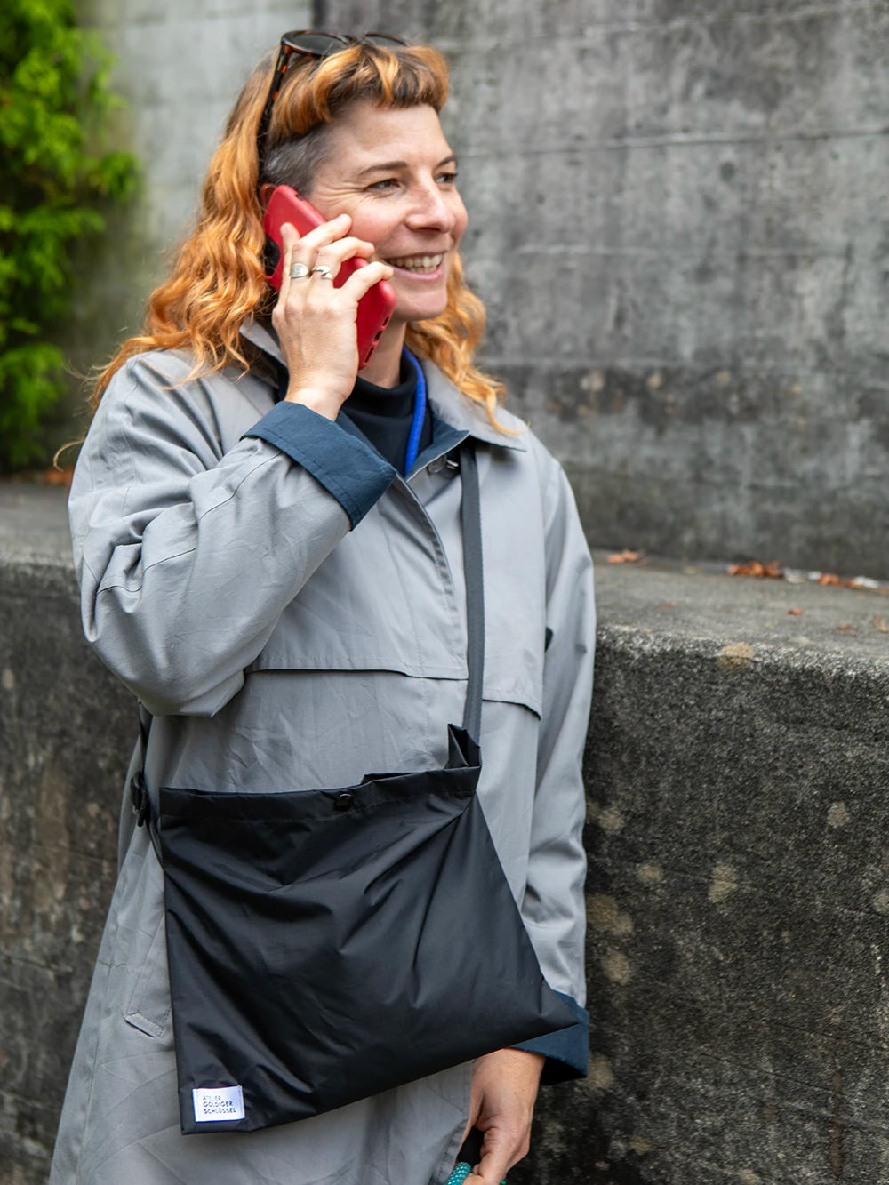 Oberkörper einer Frau mit orangen Haaren die gerade mit ihrem roten iPhone telefoniert. Sie trägt eine hellgraue Jacke und eine schwarze Umhängetasche. Im Hintergrund sieht man unscharf eine betonierte Wand.