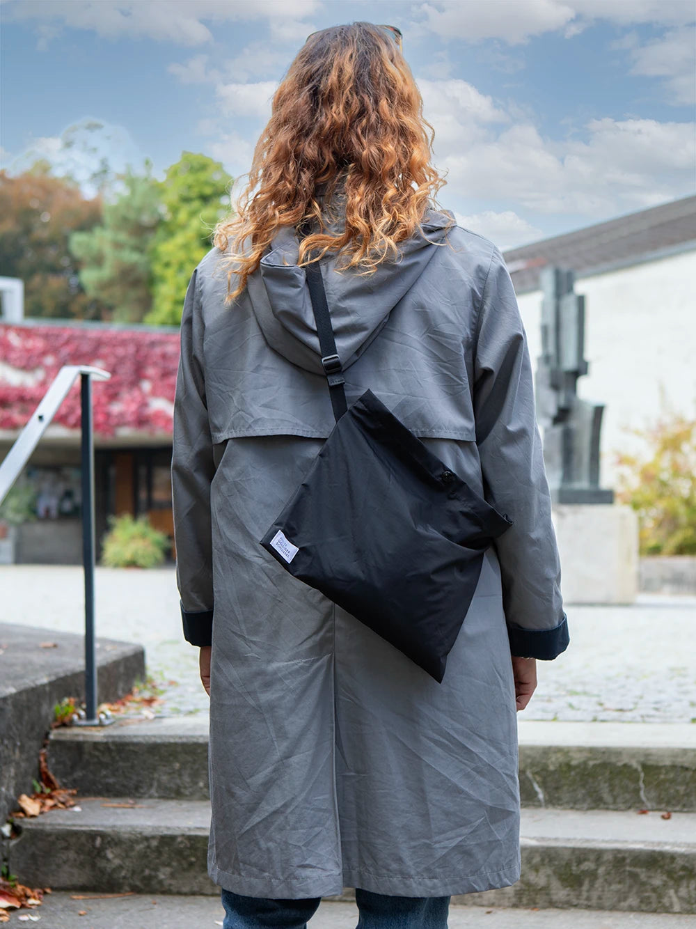 Oberkörper einer langhaarigen Person von hinten mit hellgrauer Jacke, welche eine schwarze Umhängetasche mit dem weissen Label «Atelier Goldiger Schlüssel» trägt.