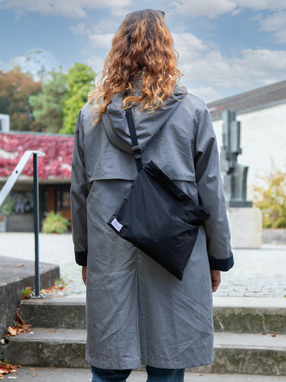 Oberkörper einer langhaarigen Person von hinten mit hellgrauer Jacke, welche eine schwarze Umhängetasche mit dem weissen Label «Atelier Goldiger Schlüssel» trägt.